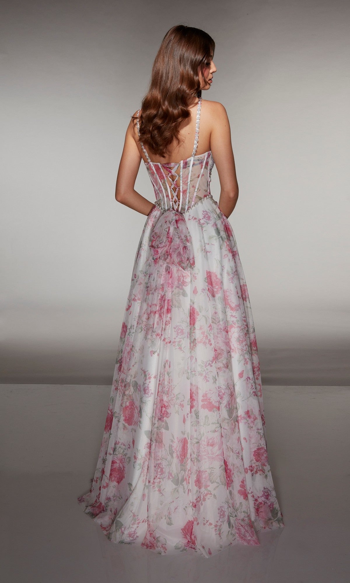 A woman with long brown hair faces away, wearing the Alyce 61994 Long Prom Dress by Alyce Paris—a stunning floral gown with a fitted corset-style bodice and thin straps.